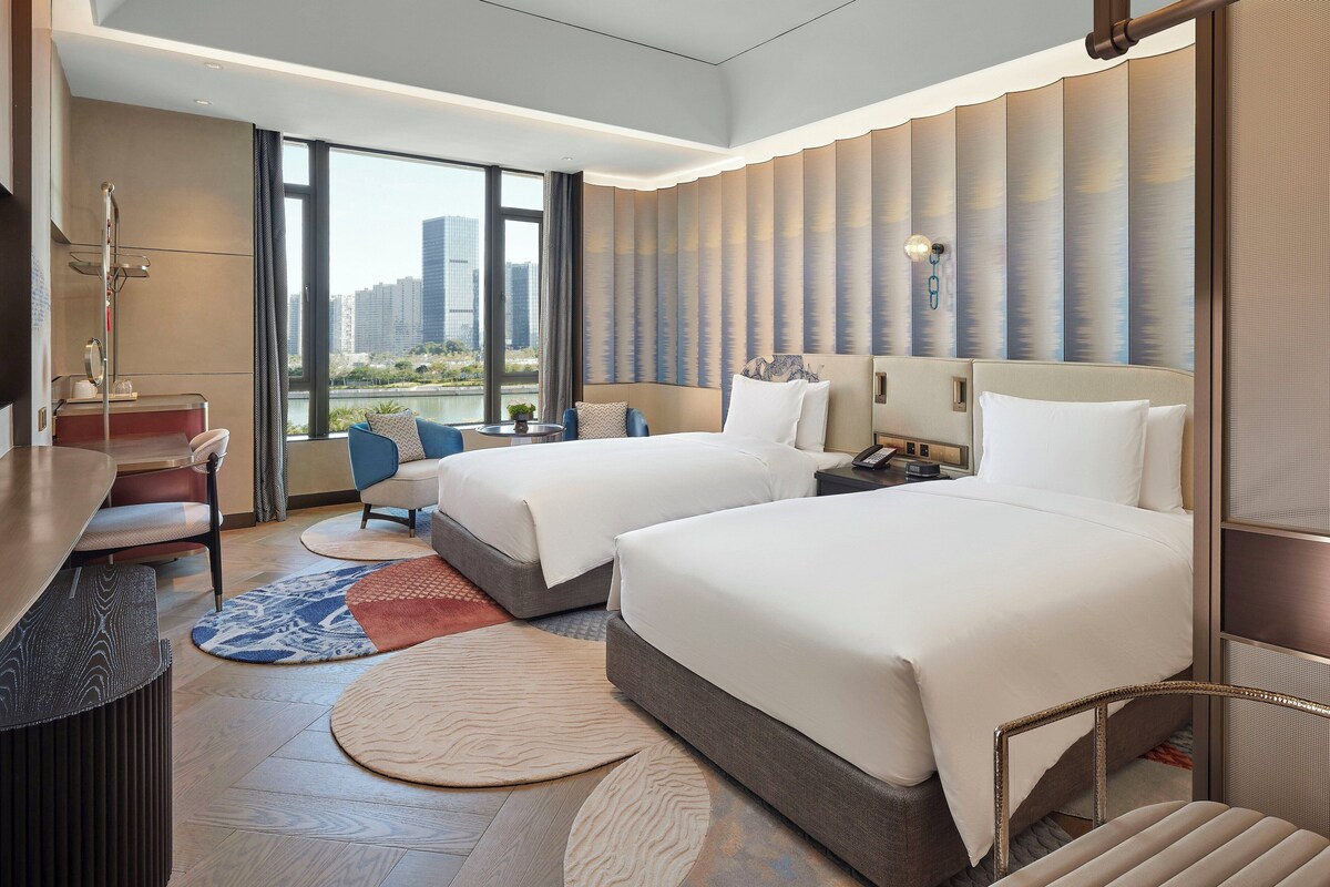 A modern hotel room with two beds, a desk, and a view of the city.