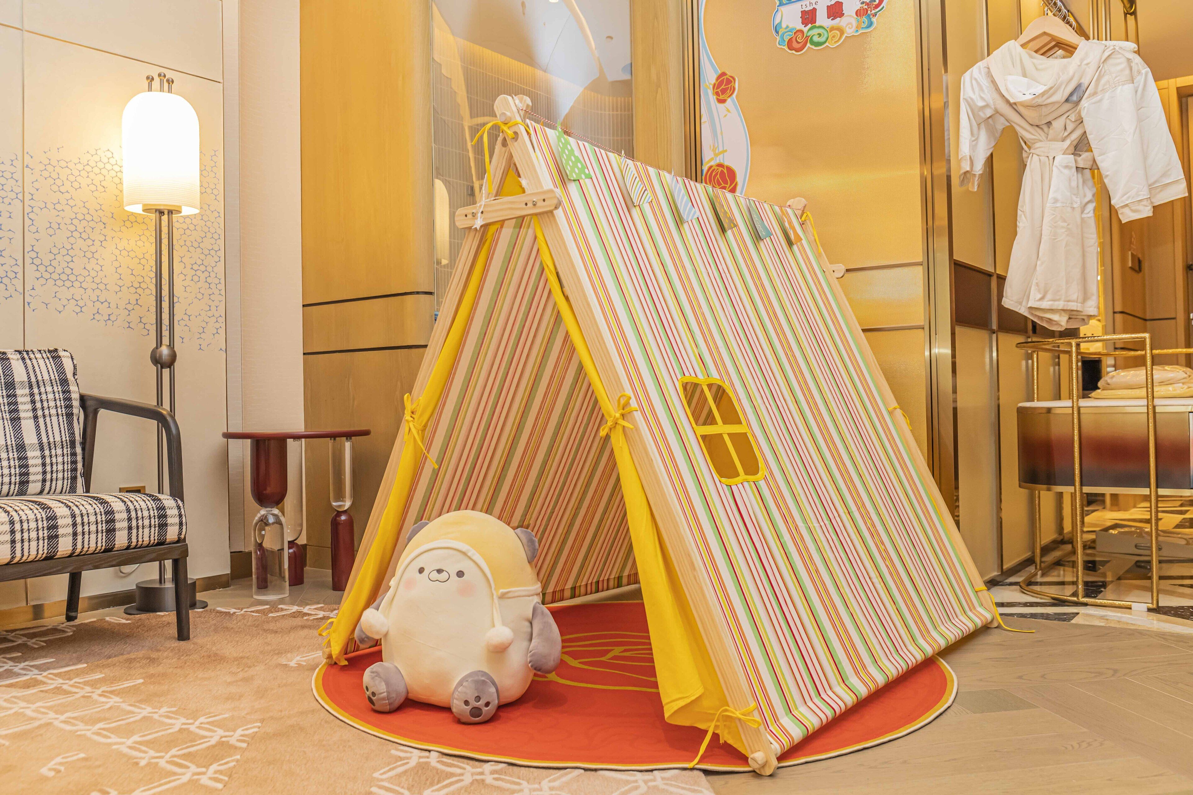 A play tent with a yellow and red striped interior, a plush toy, and a checkered chair in a hotel room.