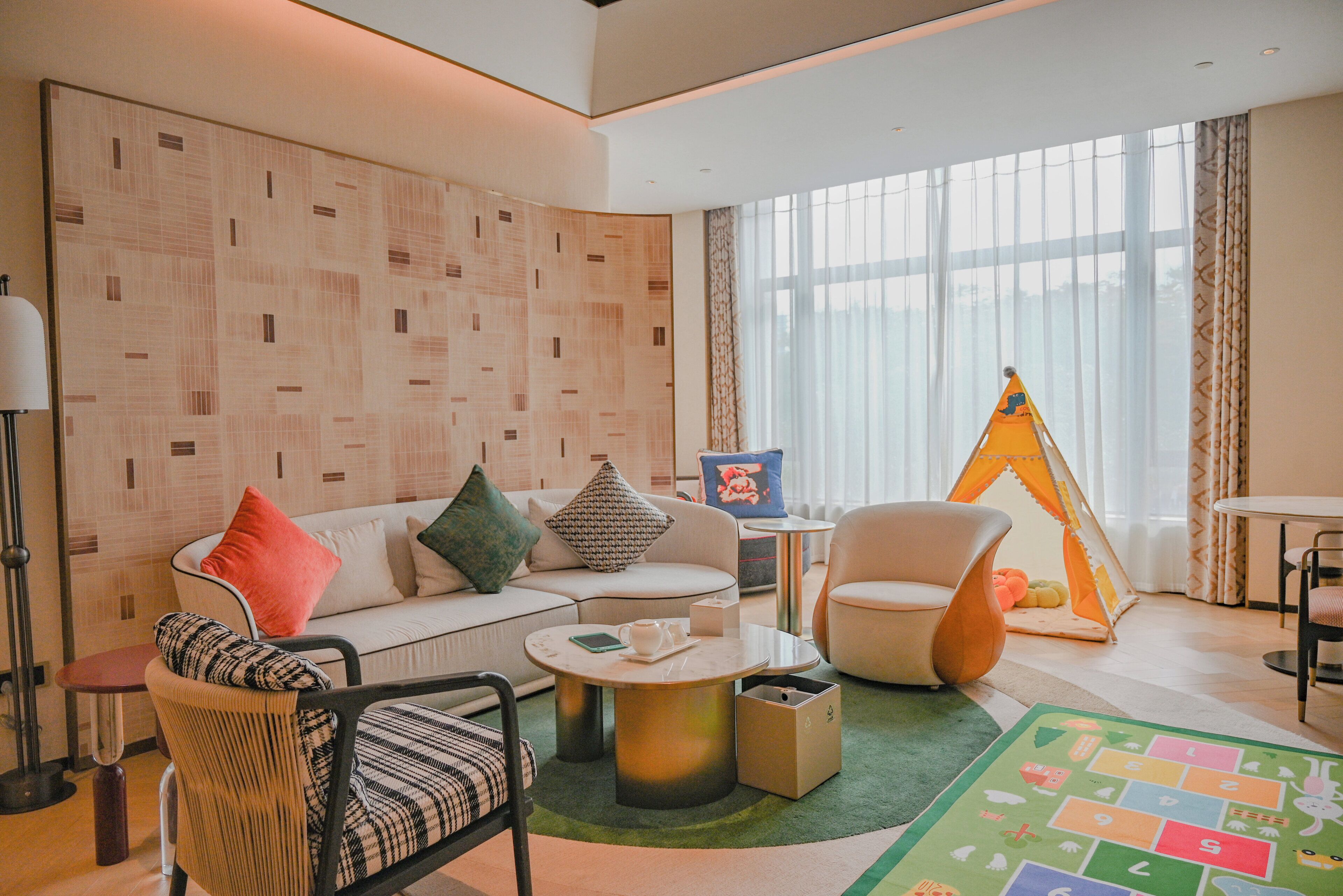A modern living room with a sofa, armchairs, a coffee table, a lamp, a tent, and a play mat.