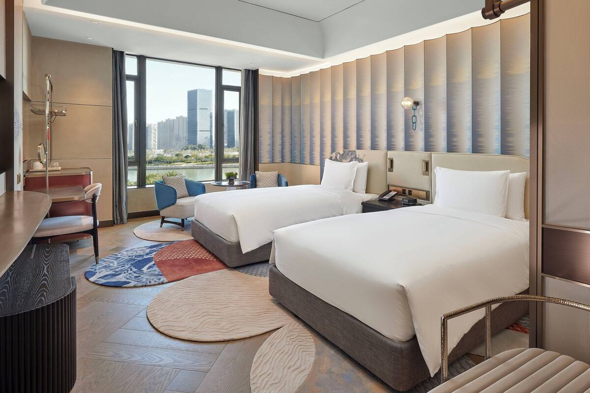 A modern hotel room with two beds, a desk, and a view of the city.