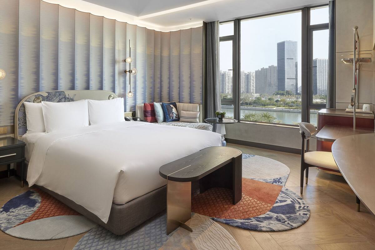 A modern hotel room with a large bed, a desk, and a view of the cityscape.