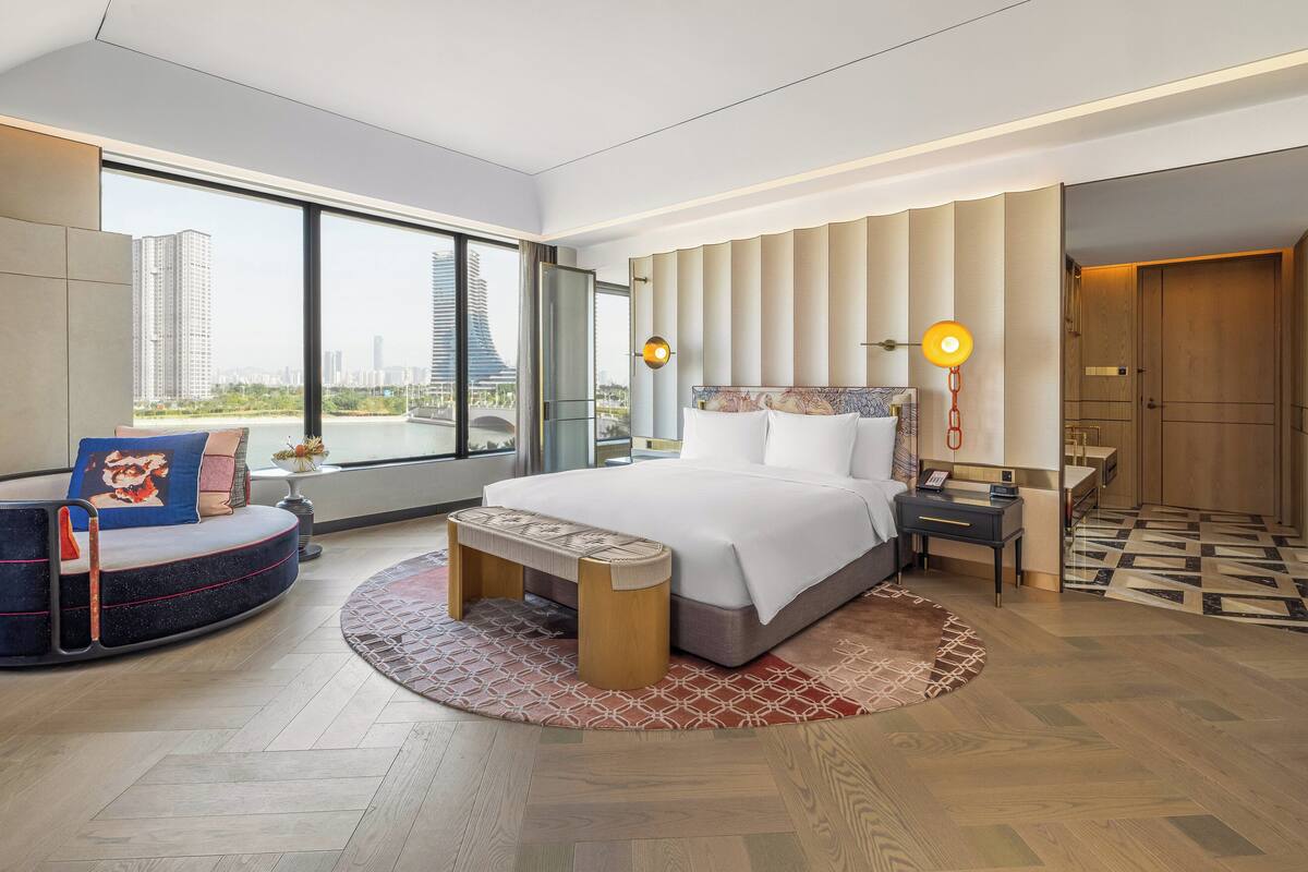 A modern hotel room with a large bed, a curved sofa, a small table, and a view of a cityscape through large windows.