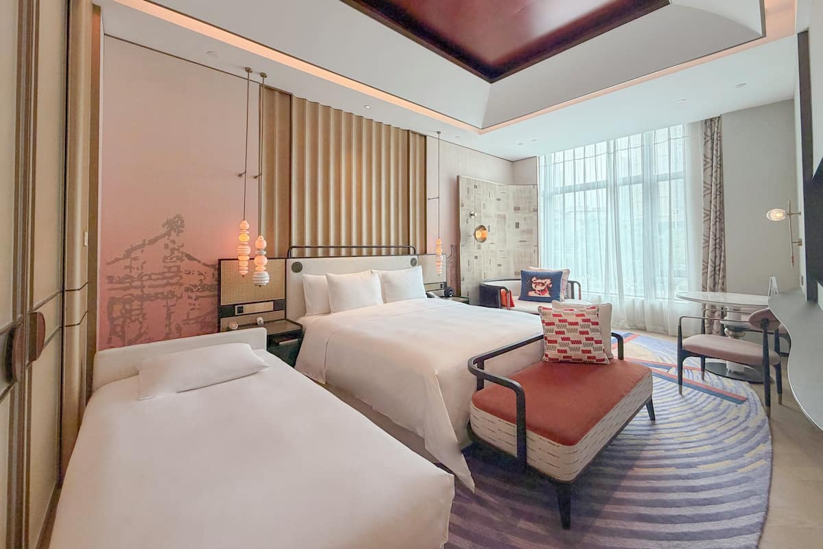 A hotel room with two beds, a desk, a chair, a large window, and a ceiling with recessed lighting.