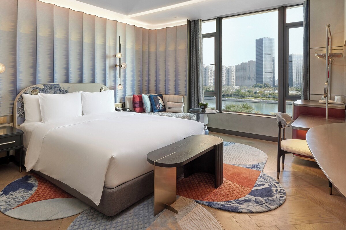 A modern hotel room with a large bed, a desk, and a view of the cityscape.