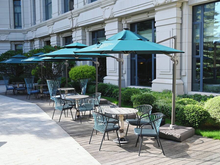 An outdoor seating area with tables and chairs, surrounded by greenery and a building facade.