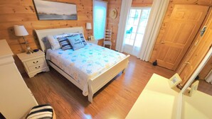 A bedroom with a wooden floor, a bed with a patterned bedspread, a nightstand, a lamp, a window with curtains, and a door. - Water View Cottage With A/C In Quiet Location, Close To Amazing Beaches (Kensington)