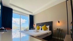 A modern hotel room with a large bed, a desk, and a view of the cityscape. - Cinematique (Mosta)