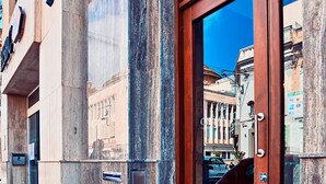A building facade with a large wooden door, a metal sign above it, and a reflection of buildings in the glass door. - Cinematique (Mosta)