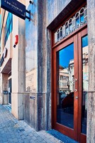 Fachada de edifício com uma grande porta de madeira, uma placa de metal acima dela e o reflexo de prédios no vidro.