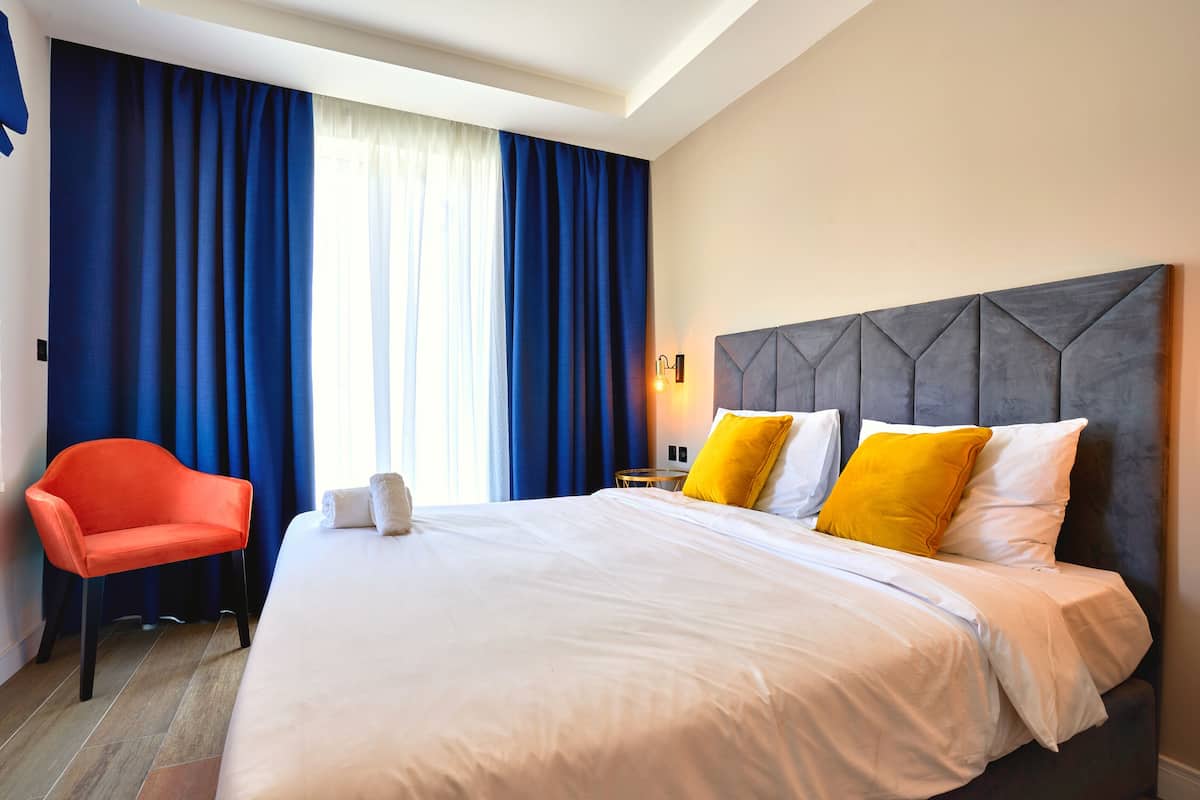A hotel room with a large bed, a bright window with blue curtains, a red chair, and a modern headboard.