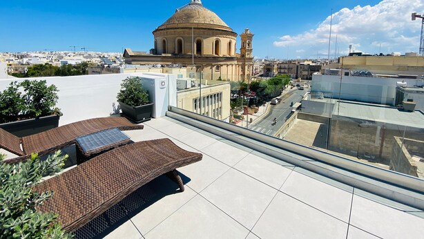 A rooftop terrace with a view of a historic building, potted plants, and a clear blue sky. - Cinematique (Mosta)