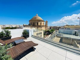 Terraço no telhado com vista para um edifício histórico, plantas em vasos e um céu azul claro.
