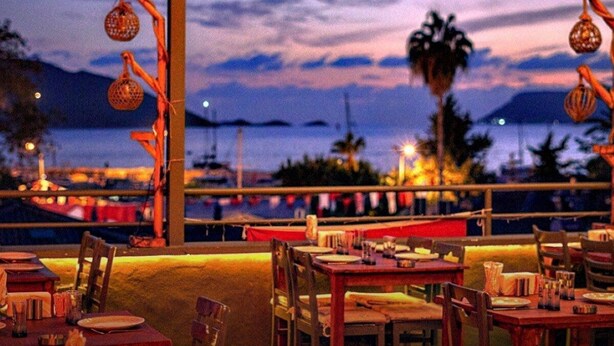 Un área de comedor al aire libre con mesas y sillas, con vistas a un puerto deportivo con barcos y un atardecer.