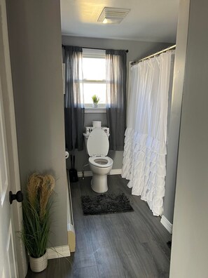 Une salle de bain avec des toilettes, une petite fenêtre avec des rideaux, un rideau de douche blanc et une plante en pot.
