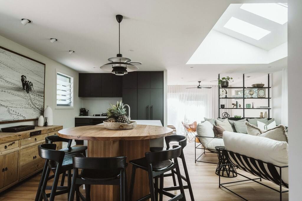 A modern kitchen with a central island, bar stools, and a dining area with a sofa and chairs. - Dunes on Sandy Wha (Gerringong)