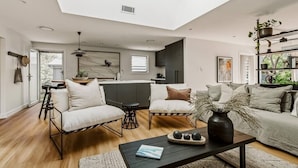 A modern living room with a skylight, a kitchen in the background, and a dining area with a table and chairs. - Dunes on Sandy Wha (Gerringong)