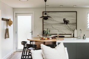 A modern kitchen with a round dining table, bar stools, a hanging light fixture, and a large abstract painting on the wall. - Dunes on Sandy Wha (Gerringong)