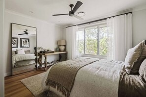 A bedroom with a bed, a ceiling fan, a window with curtains, a bedside table with a lamp, and a mirror reflecting the room. - Dunes on Sandy Wha (Gerringong)