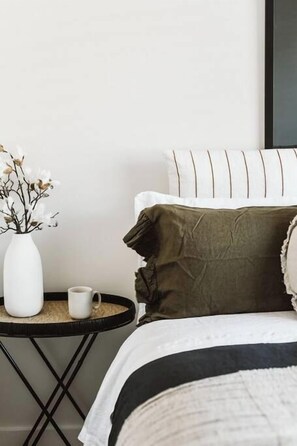 A bed with a white headboard, a green pillow, and a white vase with flowers on a small round table beside it. - Dunes on Sandy Wha (Gerringong)