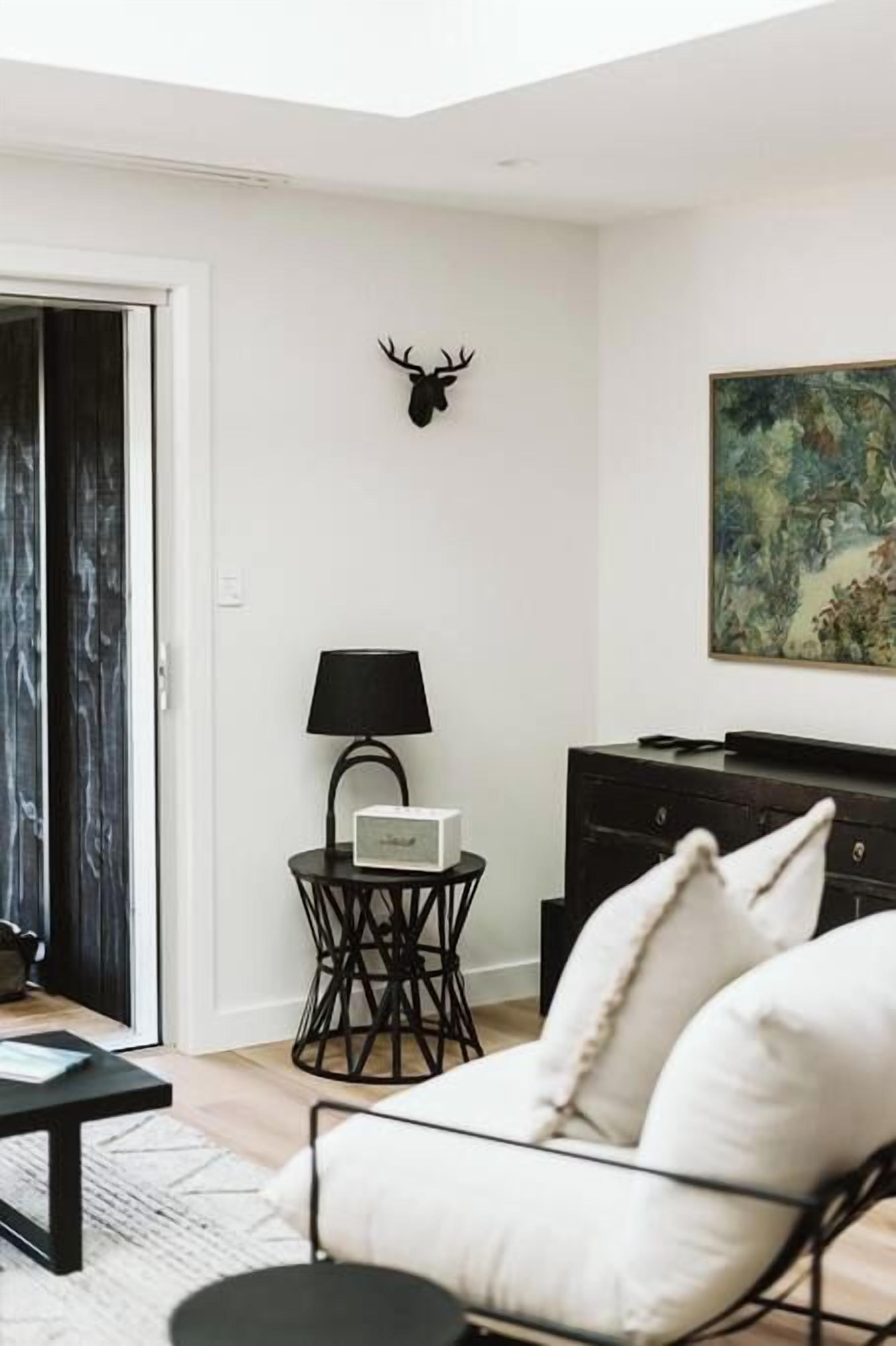 A modern living room with a white sofa, a black coffee table, a side table with a lamp, a black cabinet, and a deer head mounted on the wall.