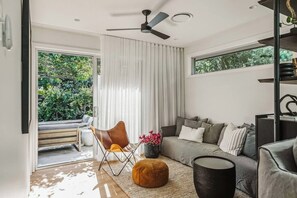 A modern living room with a grey sofa, a brown leather chair, a round ottoman, and a ceiling fan. - Dunes on Sandy Wha (Gerringong)
