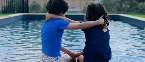 Dos niños sentados junto a una piscina, con una casa al fondo.