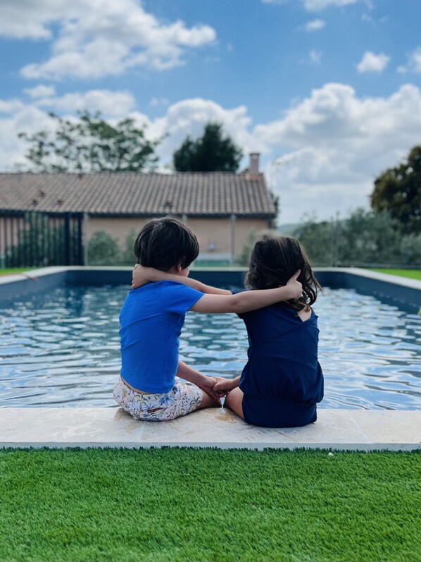 Two children sitting by a swimming pool with a house in the background. - Gîtes 3 chambres -Piscine -Vue sur la vallée de l'Orb (Cessenon sur Orb)