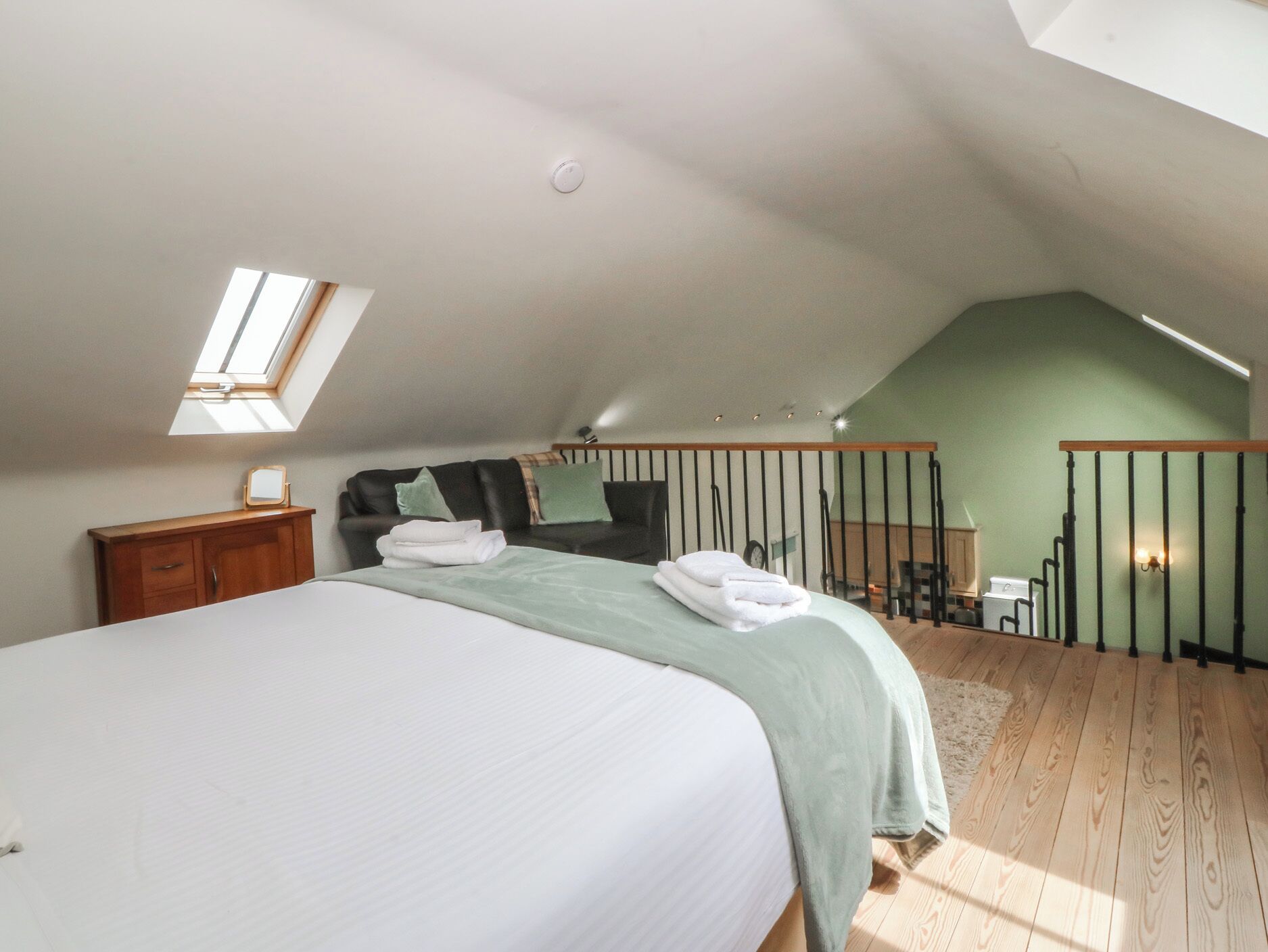 Une chambre à coucher avec un lit, une table de chevet, une commode en bois, un mur vert, un plancher en bois et un puits de lumière.