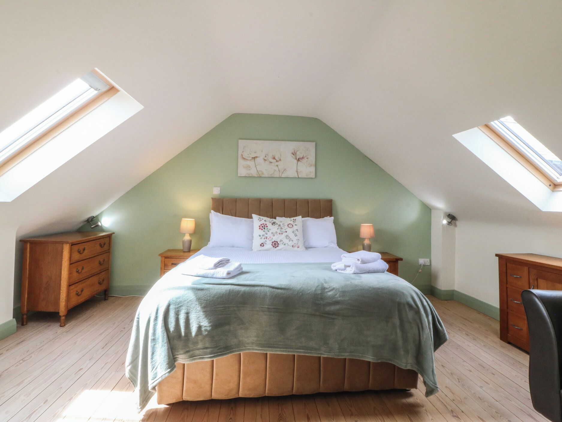 Une chambre avec un plafond en pente, un lit avec des oreillers et une couverture, deux tables de chevet avec des lampes et un tableau au mur.