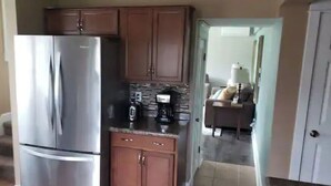 Uma cozinha com geladeira de aço inoxidável, armários de madeira, bancada com cafeteira e vista para uma sala de estar.