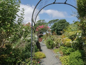 Een tuinpad met een bakstenen boog, omgeven door verschillende planten en bloemen.