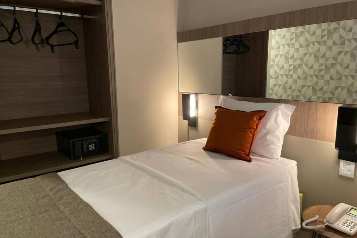 Habitación de hotel con cama, mesita de noche con teléfono, espejo y armario con perchas, además de un diseño geométrico en la pared.
