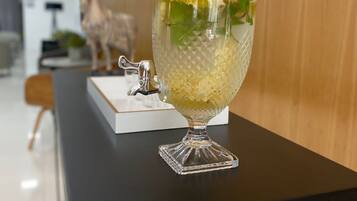 A clear glass punch bowl with a lid, filled with a yellowish liquid, lemon slices, and mint leaves on a black table.