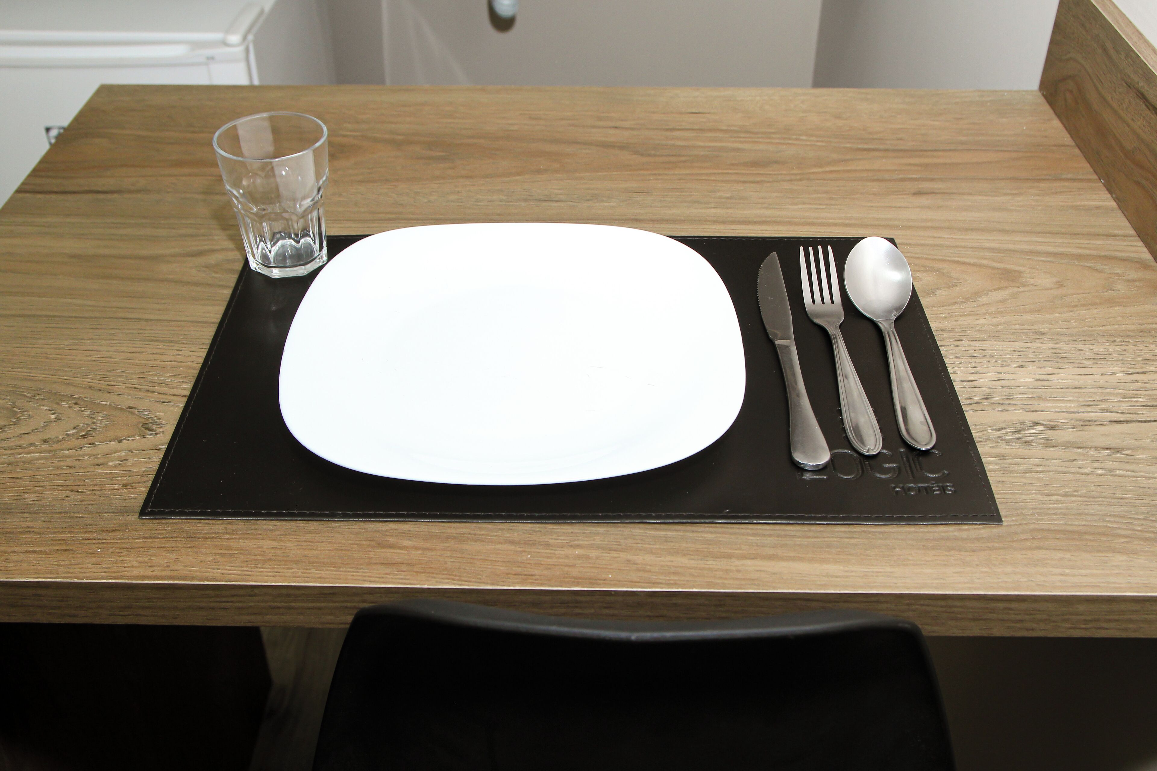 A wooden table with a black placemat, a white plate, and a set of cutlery including a knife, fork, and spoon.