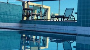 Una piscina azul cristalina con bordes de azulejos, barandilla de vidrio y dos sillas de descanso bajo una sombrilla.