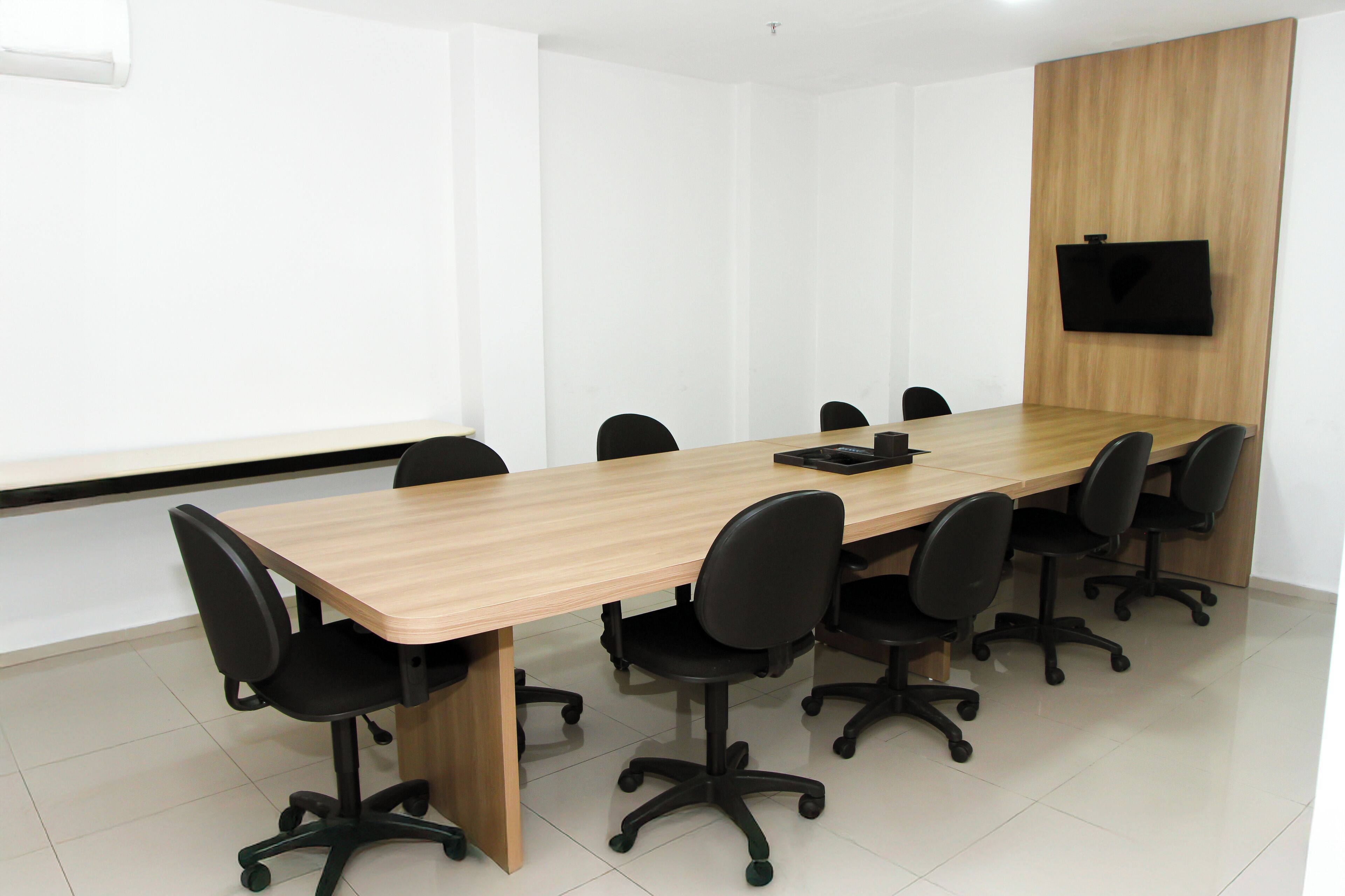 A conference room with a long table, black office chairs, and a flat-screen TV mounted on the wall.
