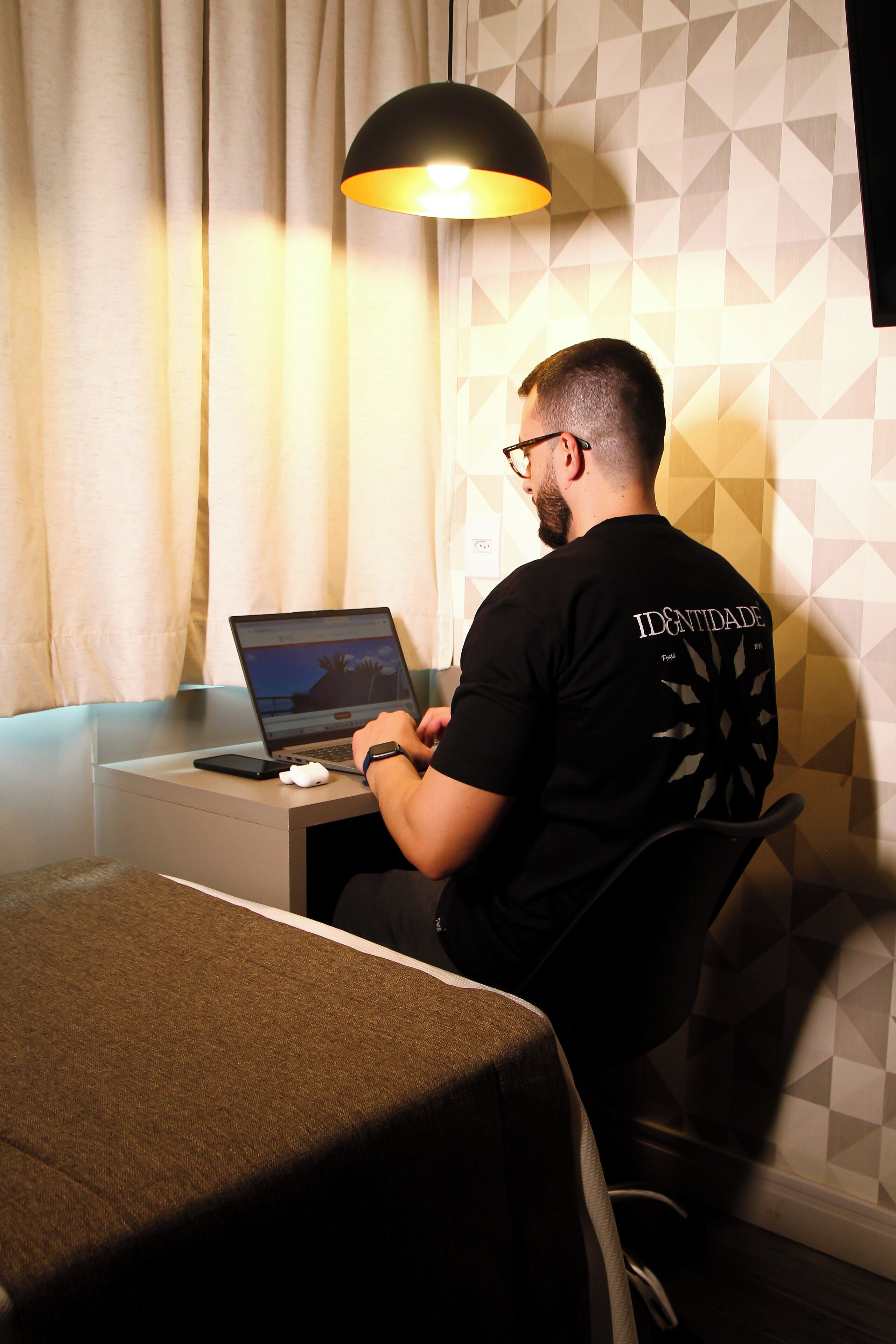 A person sitting at a desk with a laptop, a smartphone, and a mouse, in a room with a geometric patterned wall and a curtain.
