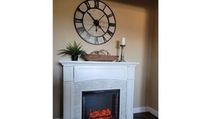 Une cheminée avec un manteau de couleur blanche, une grande horloge murale, une plante en pot et un chandelier.