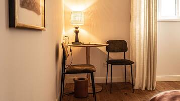 Una habitación de esquina con piso de madera, una mesa pequeña con lámpara, una silla y un cuadro enmarcado en la pared.