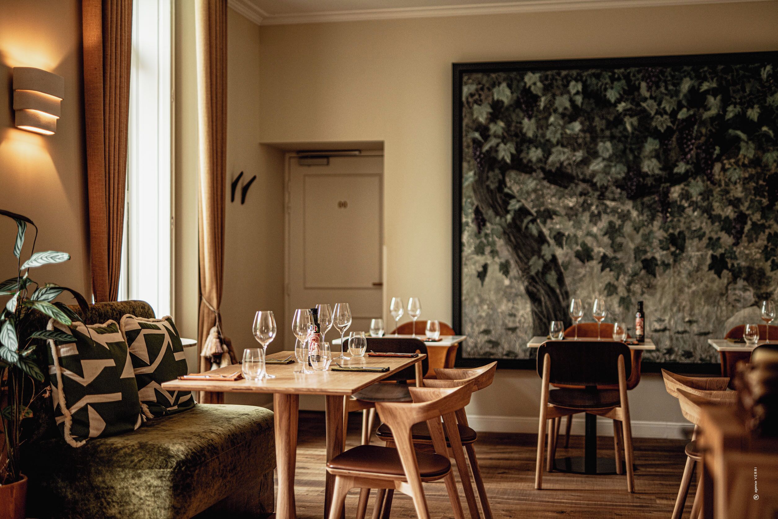 A dining area with wooden tables and chairs, a sofa with patterned cushions, and a large floral painting on the wall.