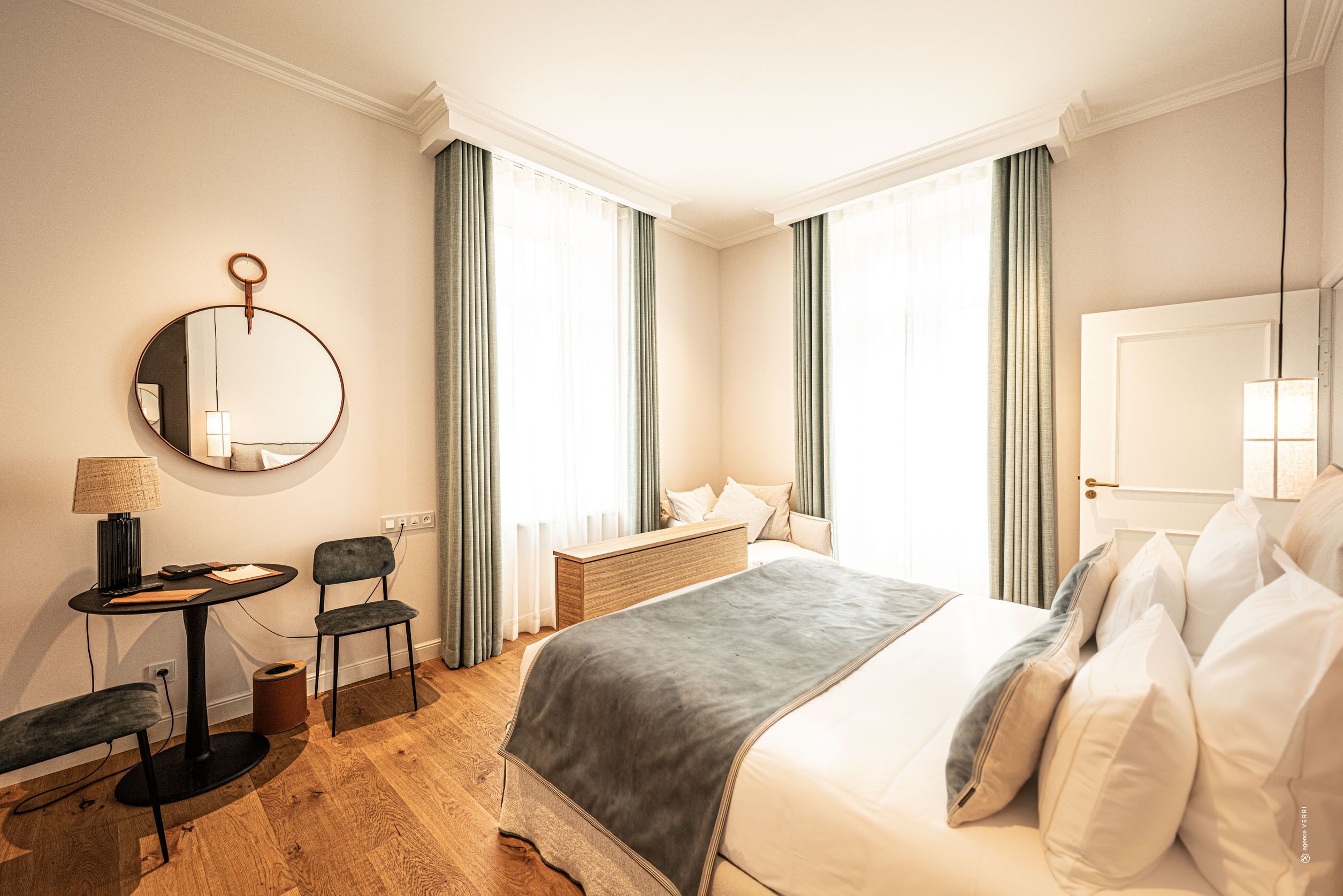 A hotel room with a bed, a desk with a lamp, a chair, a round mirror, and a window with curtains.