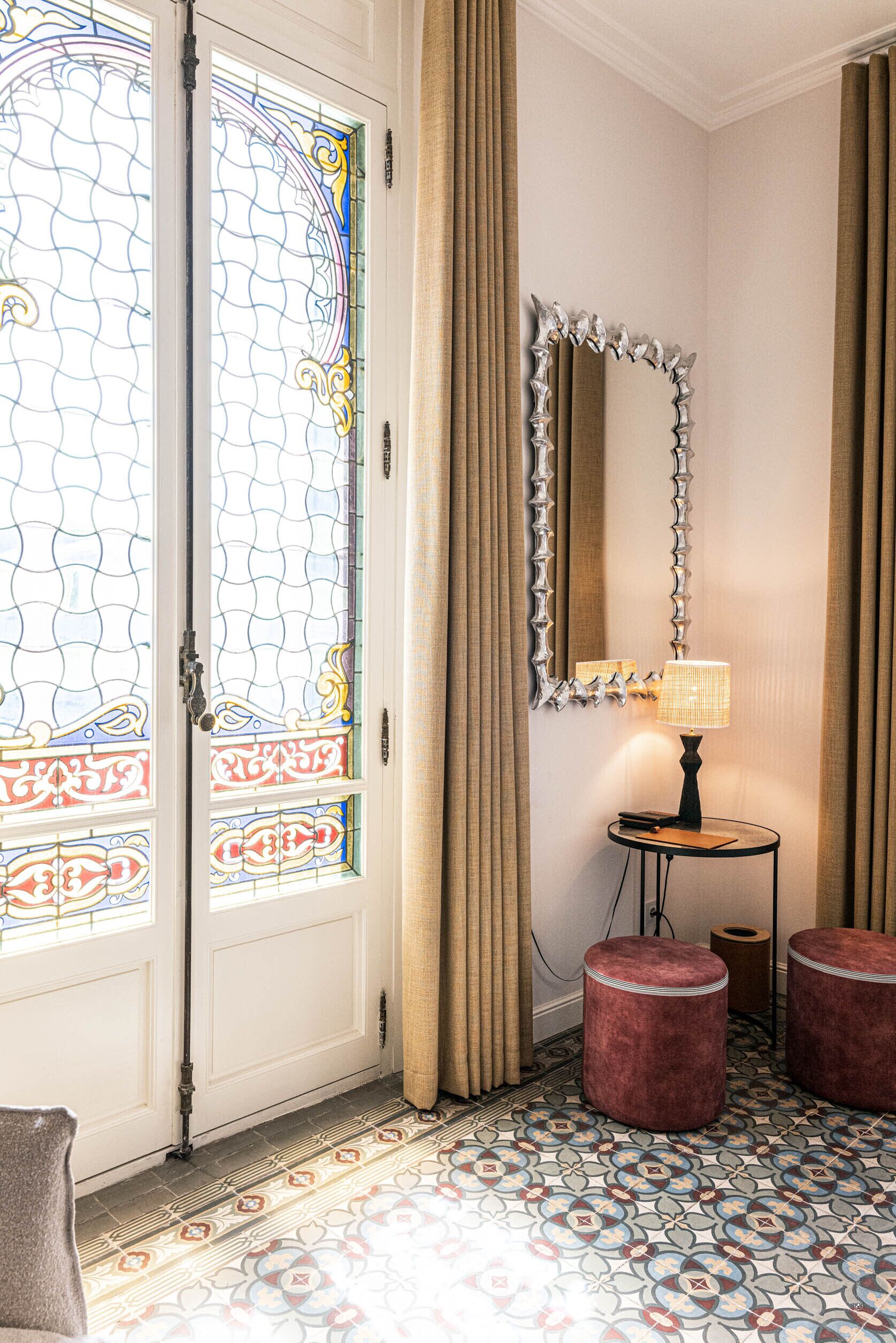 A room with a patterned floor, a door with stained glass, a mirror with a decorative frame, and a table with a lamp.
