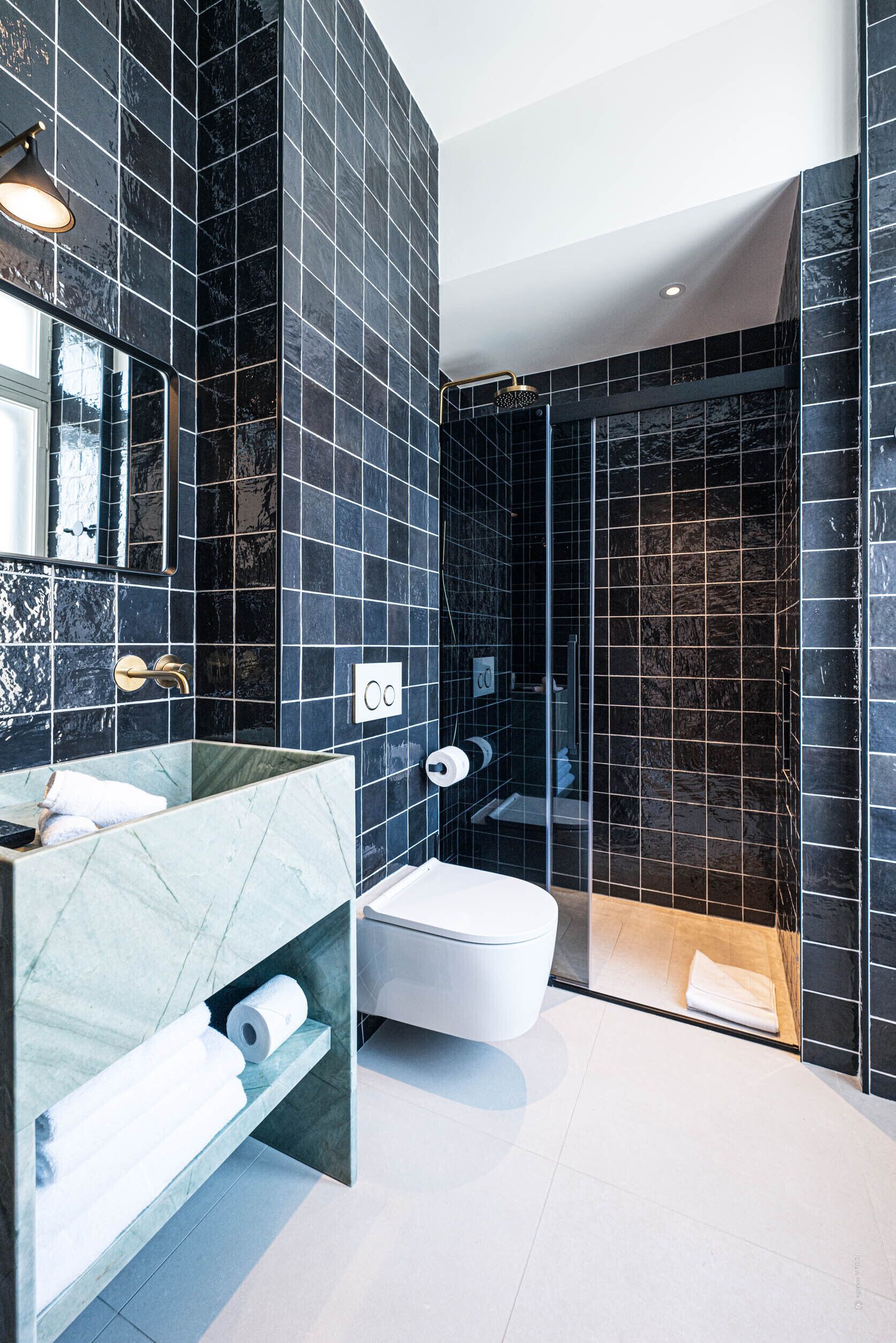Un baño moderno con paredes de azulejos oscuros, un tocador de mármol, un inodoro blanco y una zona de ducha con mampara de vidrio.