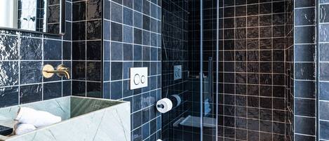Un baño moderno con paredes de azulejos oscuros, un tocador de mármol, un inodoro blanco y una zona de ducha con mampara de vidrio.