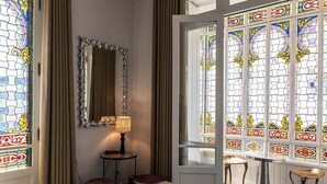 Een kamer met een houten receptiebalie, een spiegel, een lamp, een klein tafeltje met krukjes, en een groot glas-in-lood raam.