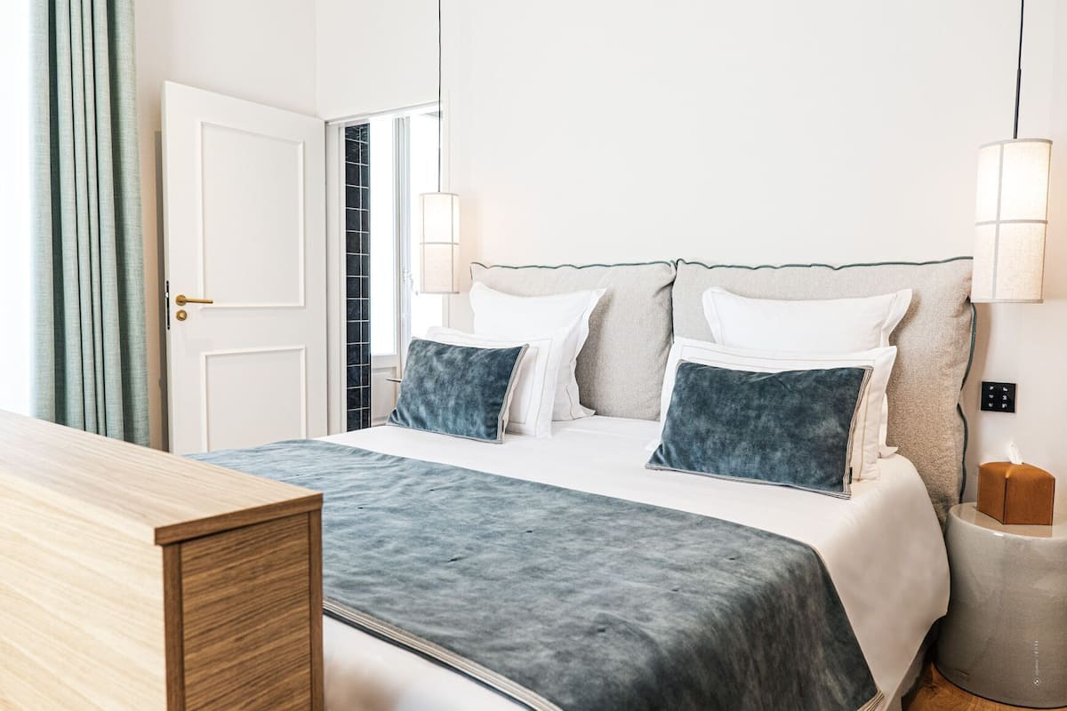 Een opgemaakt bed met een blauw-witte dekbedovertrek, een nachtkastje, een lamp en een nachttafeltje met een zak tissues.