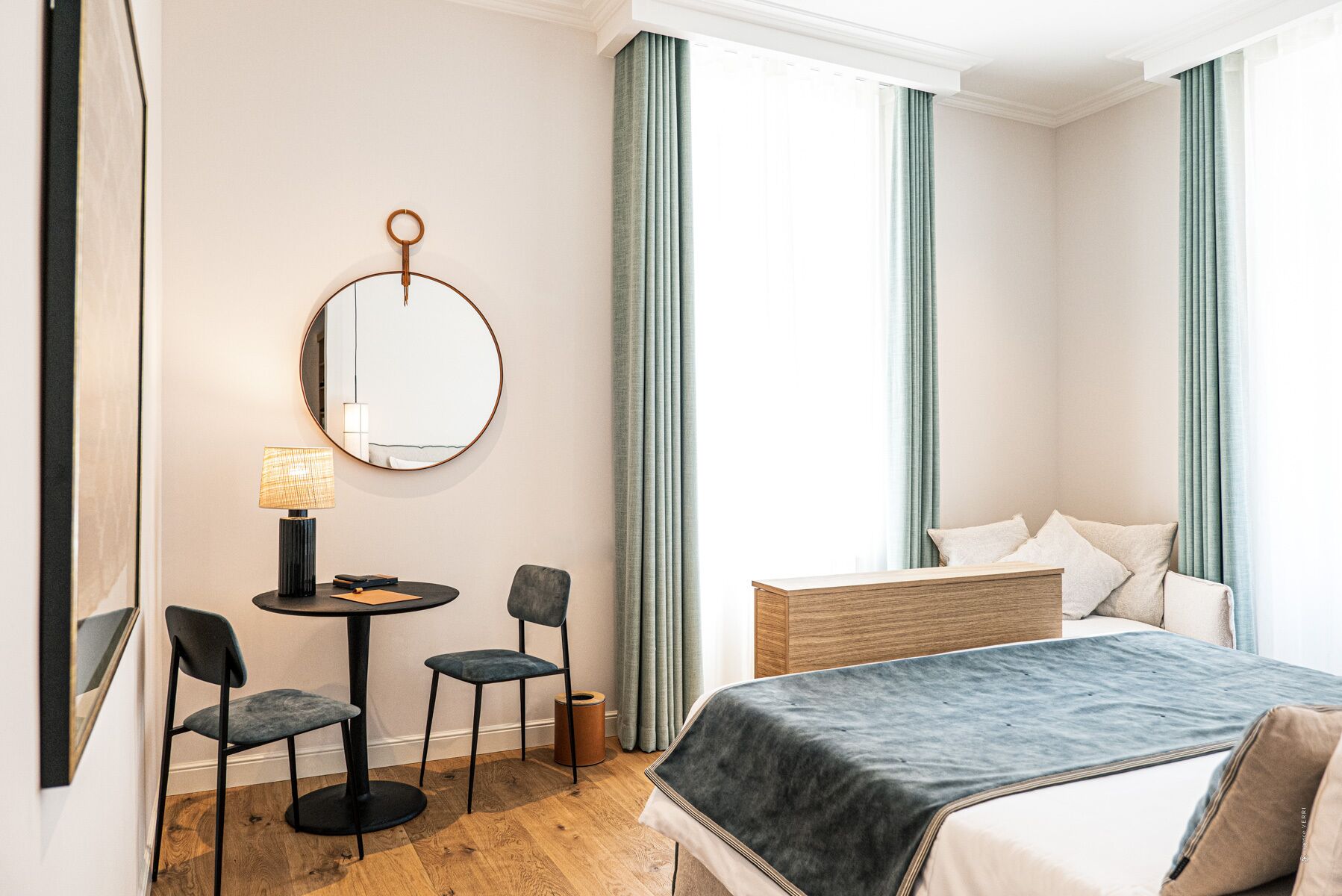 Une chambre d’hôtel avec un lit, un bureau avec deux chaises, un miroir rond et une lampe.