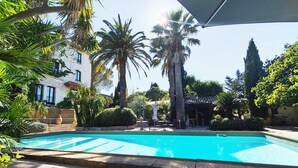A swimming pool with a tiled deck, surrounded by palm trees and a building with a tiled roof. - 2 -Studio Lierre Les Bastidières (Toulon)