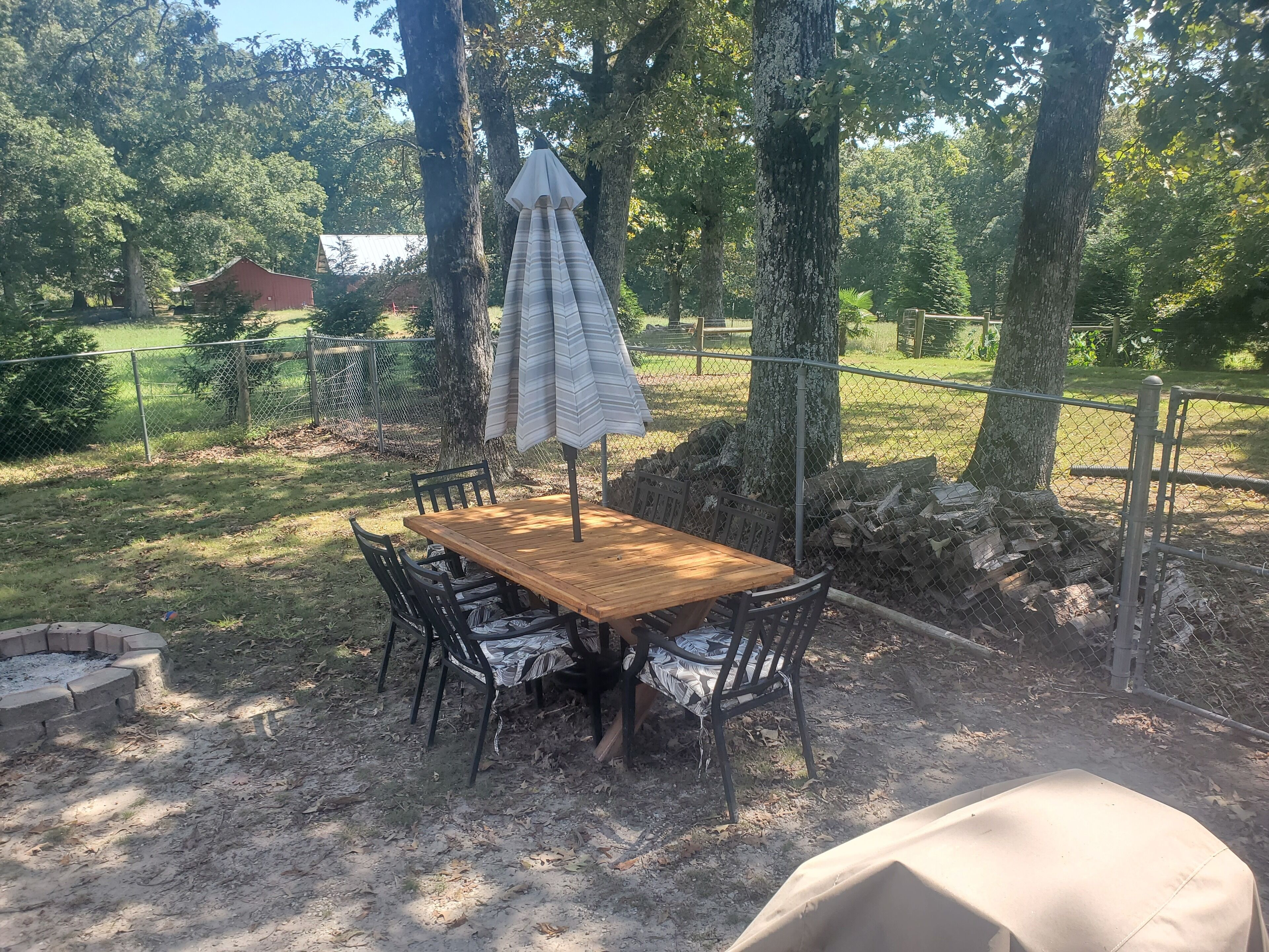An outdoor dining area with a wooden table and chairs, surrounded by trees and a chain-link fence.