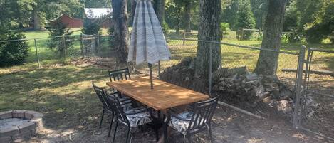 An outdoor dining area with a wooden table and chairs, surrounded by trees and a chain-link fence.
